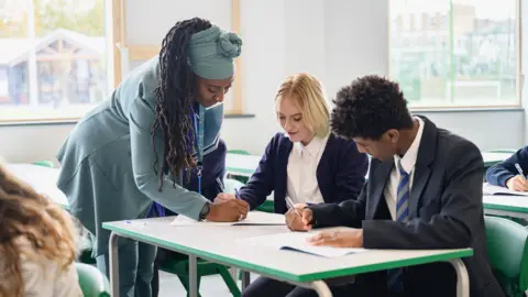 Getty Images A female teacher with long braids in a pale blue coat with a matching blue headwrap, helping a blonde student in a white shirt and a male student with a white shirt and blue striped tie their school work. Sat in a white classroom with windows and green chairs.