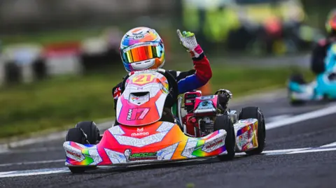 Deno holds a finger in the air as he drives a brightly coloured kart and wears a matching driver kit and reflective helmet
