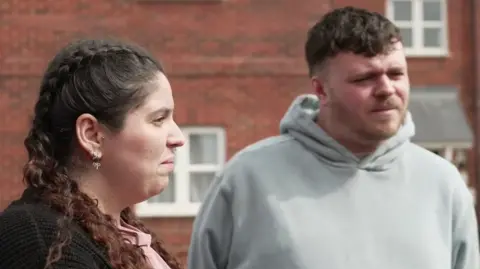 Samantha and Joshua stand next to each other outside their flat. They are looking at someone just off camera.