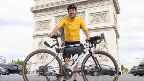 PA Chris Boardman in a yellow cycling jersey stands in front of the Arc de Triomphe in Paris.
