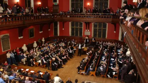 PA Media A view from the gallery of the packed debating chamber which has red walls and large windows. The young man speaking on the floor of the room is wearing evening dress and those sitting near him are similarly attired. Audience members and those in the gallery wear less formal clothes.