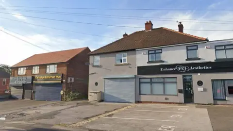 A row of shop units in a residential street. The unit in the centre has metal shutters down and is unused.