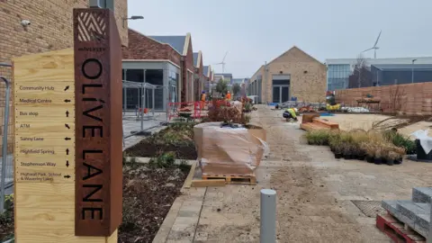 A sign saying 'Olive Lane' together with a list of facilities. Construction work and equipment in the background with people in hard hats behind site fencing