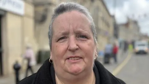 A woman with short cropped grey hair. She is wearing a black coat and is standing in the street. She looks at the camera with a neutral expression.