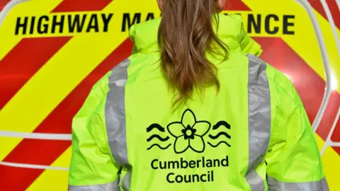 A Cumberland council worker facing a Highway Maintenance vehicle. Her back is turned to the camera. She has a long brown hair in a ponytail and wears a high visibility jacket with the council logo.