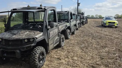 A row of five grey John Deere Gators and a telehandler lined up bumper to bumper on the edge of a ploughed field. A police car is parked to the right of the frame.