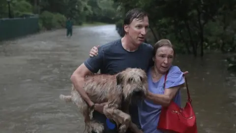 Getty Images A man helps a neighbour down a street in Houston after rescuing her from her home in his boat in River Oaks, Houston (27 August 2017)