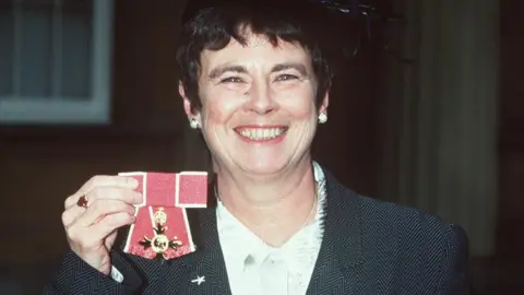 BBC News angela mason holds up her oBE while she smiles at the camera