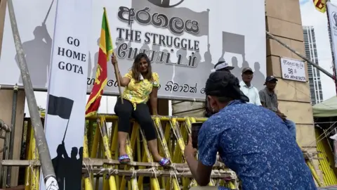 BBC/Tessa Wong Man snaps picture of a woman posing on protest village stage