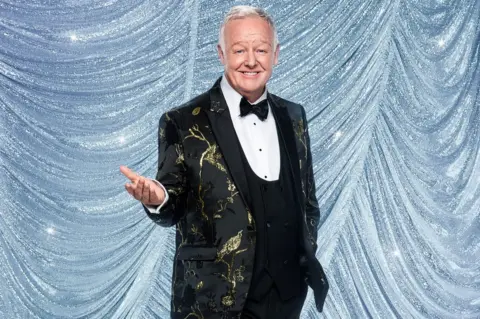 Ray Burmiston/BBC Les Dennis with his palm face up wearing a black and gold jacket