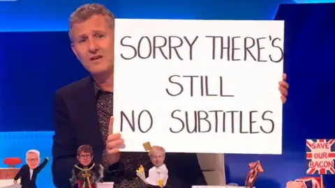 Channel 4 Adam Hills on Channel 4's The Last Leg holding up a sign reading "Sorry there's still no subtitles"