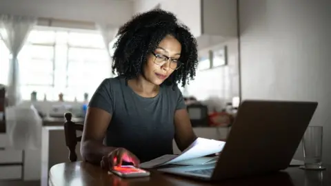 Getty Images Woman looks at bills while holding a smartphone in front of a laptop