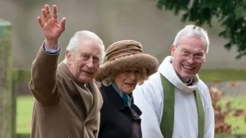 PA Media King Charles III waves to the cameras as he walks beside Queen Camilla