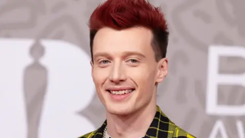 Getty Images Jack Saunders attends the 2023 Brit Awards. Jack is a 31-year-old white man with dyed red hair that is long on top. He has blue eyes and smiles at the camera from the red carpet. He wears a yellow and black tartan suit