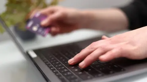 PA Media A woman using a laptop as she holds a bank card