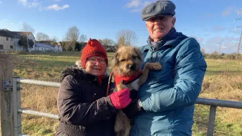 BBC Leila and Mike standing looking at the camera holding their dog between them in winter clothes, Leila in a red woolly hat, Mike in a flat cap in front of a field