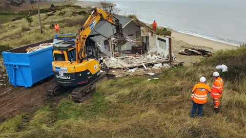 Shaun Whitmore/BBC A house is partially demolished next to the cliff edge with a bright yellow digger next to a blue skip.