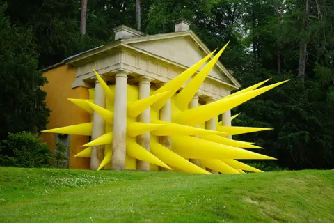 Christopher Furlong / Getty Images A yellow inflatable artwork with spikes is seen bursting through the pillars of the Temple of Piety at Studley Royal