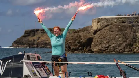 World's Toughest Row Leanne standing on her boat on arrival in Antigua, holding two red flares in the air in celebration.