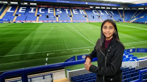 Nigel Maitland Renuka at Leicester City's stadium. Behind her is the green pitch and empty stands with blue seats, with white writing spelling out Leicester City. Renuka is looking at the camera, leaning on blue railings, with her left hand over her right hand. She is wearing a black jacket with a maroon turtle neck top.