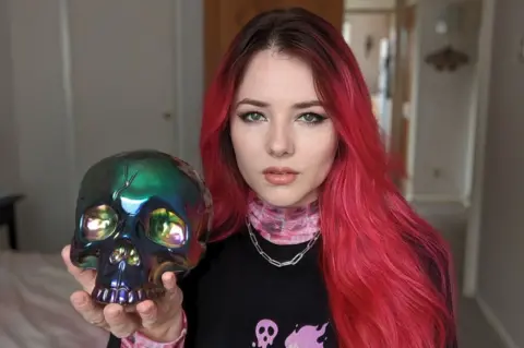 Ash Millman A woman with long, red/pink hair sits on a bed, holding an iridescent skull in her upturned left palm. She's giving a piercing stare to the camera, and wears a black top with pink-pattered high neck. She's got a chain-style silver necklace on too.