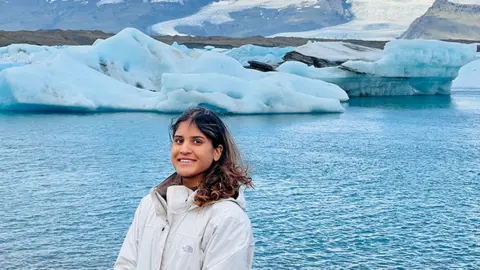 Serena Sahota Serena posing for a photo with the sea and some white glaziers visible behind her as well as mountains. She is smiling and her dark brunette hair looks a bit windswept over her shoulders. She is wearing a white coat with the hood down and it is zipped up.