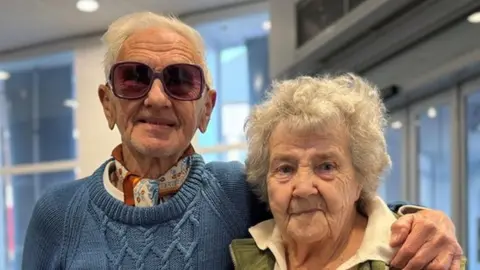 Man with short white hair and dark glasses wearing a blue sweater has his arm round a woman wearing a green gilet over a white shirt