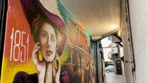 Katy Lewis/BBC The colourful mural, featuring the date 1851 and a Victorian woman's face, in an alleyway