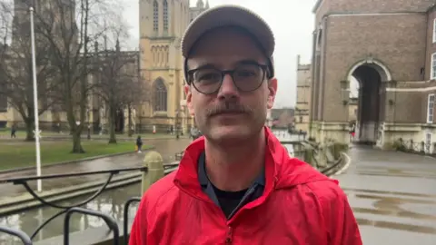 Sam Kidel in a red Acorn branded jacket and beige cap, wearing black thick-rimmed glasses