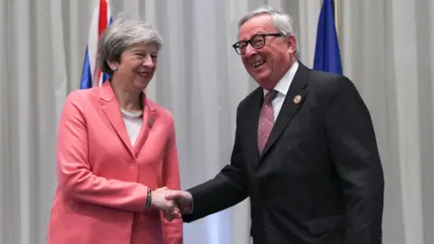 AFP Theresa May and Jean-Claude Juncker smile and shake hands in front of the flags of their respective nations