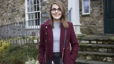 Williams family Elin wearing a coat and sweater standing in front of a building with traditional style stone wall