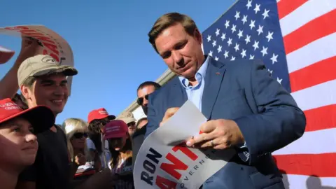 Getty Images Ron DeSantis signs a poster at a 2018 campaign event with a US flag in the background