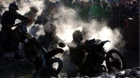 Three motorbike riders covered in mud and dust watched by spectators