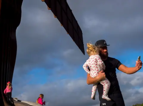 Peter Dench A man takes a seflie on his phone holding a young girl in front of The Angel of the North, a contemporary sculpture, designed by Antony Gormley. Gateshead, Tyne and Wear.