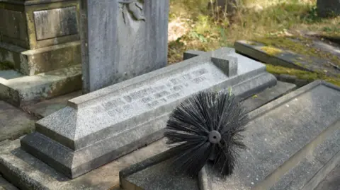Trafford Council A grey headstone with a chimney sweep brush laid on it.