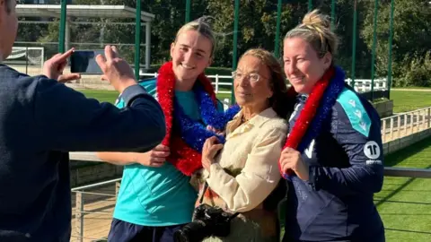 Hy Money, tenant un appareil photo, se tient entre deux femmes souriantes en tenue de sport, drapées de guirlandes rouges et bleues. Un homme prend sa photo en plein air sur un terrain de sport.