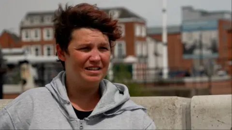Becki Bowden/BBC A woman with short brown hair and wearing a grey hoodie stands next to Hull Marina. There are several large buildings behind her.