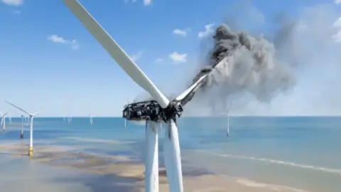 Olivers Drone Photography Smoke is emitting from the right-most turbine blade, with the blackened hub and turbine generator housing. Beyond are other wind turbines, unaffected, and the sea. The turbines in the near ground are on a sandbank which is coloured yellow and brown.