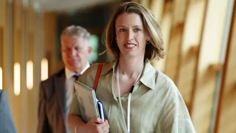 PA Media Hannah Mary Goodlad walking in a corridor at Holyrood. She has short blonde hair and is smiling while carrying a folder under her arm.