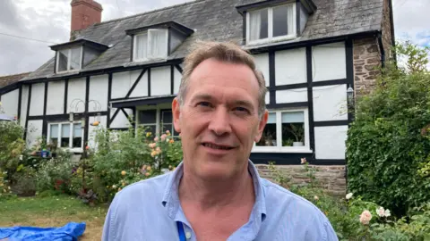 Dominic Harbour has short brown hair and is clean shaven. He is wearing a light blue shirt and is stood in front of a black and white cottage.