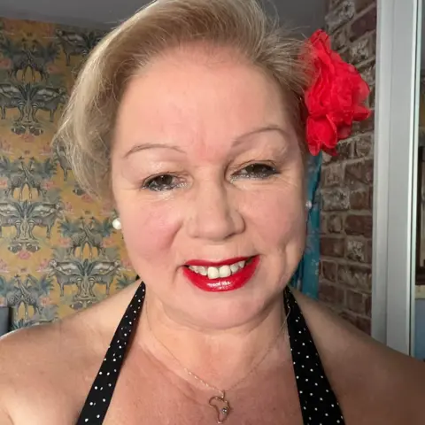 Mandy Polkey Selfie of a smiling Mandy Polkey who is dressed in 1950s style with a black halter-neck top with white polka-dots. She has blonde hair worn up with a red flower on the right side. She is also wearing red lipstick.