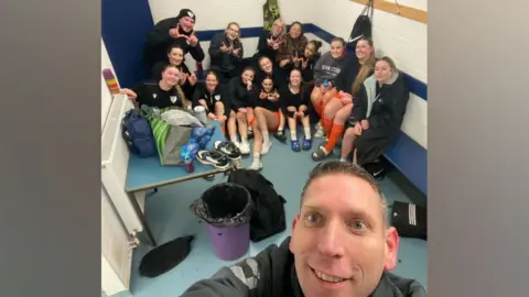 A selfie style photo with the club assistant coach at the bottom smiling at the camera and the girls behind. The team are in a locker room