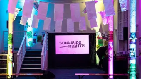 Rhiannon Banks Sunniside Nights writing is projected on a screen. Light rectangular flags hang between pillars covered in flyers. Projectors light up the room in blue, pink and yellow.
