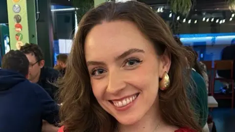 PETRA WELLS Petra smiling at the camera. She has long brown hair and is wearing gold earrings.