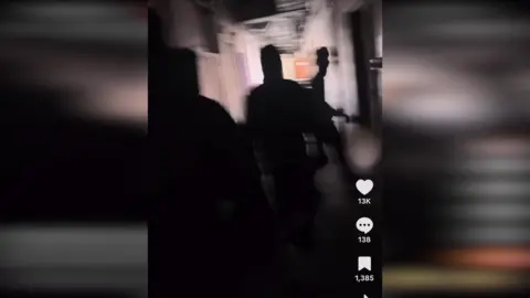 TikTok A still from a Tiktok video showing the back of several people walking along a hallway. They are obscured from view and appear all in black. 