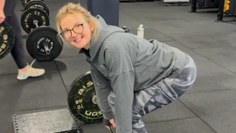 Victoria Skinner Tor bending down to lift a weight, wearing a grey jumper and sport leggings, with blond hair tied back and glasses in. She is in a gym. 