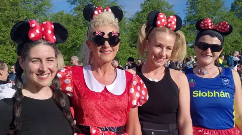 Four women standing next to each other. From left to right the first has plaited long brown hair, black top and is wearing Minnie Mouse ears. Next to her is Elaine who has blonde hair tied back wearing a red Minnie Mouse costume. Next to her is a lady with black clothing and short blonde hair wearing Minnie Mouse ears, and beside her is a lady with a blue top, red shorts black sunglasses and Minnie Mouse ears with her dark hair tied up.