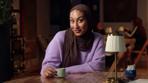 Mariah is wearing a brown headscarf over a lilac jumper while holding a small cup on a saucer. She is in a cafe with some people sat at a table slightly blurred out in the background. She is smirking in the image.