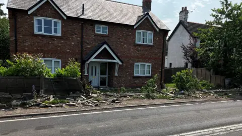 BBC A detached two-storey house set back from a main road. The brick wall and fence in front of it has been destroyed, with rubble lying everywhere.
