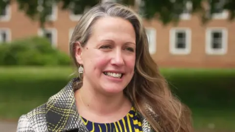 Councillor Cheryl Cottle-Hunkin smiling and looking to the right. She has long brown hair and is wearing a yellow and grey coat with a blue and yellow top.